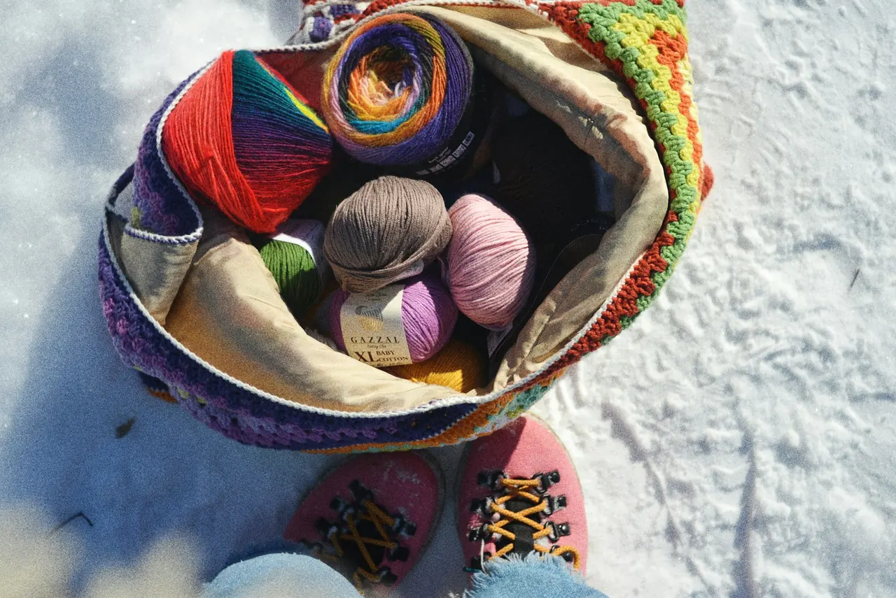 An open tote of colorful yarn cakes resting on snow beside pink hiking boots