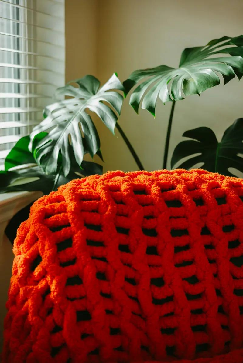 A chunky orange knitted throw in front of a monstera plant and window blinds